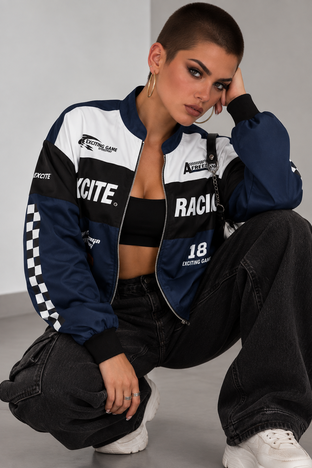 A model wearing a navy blue, white, and black racing-themed bomber jacket, paired with black cargo pants and white sneakers, posing in a studio setting.
